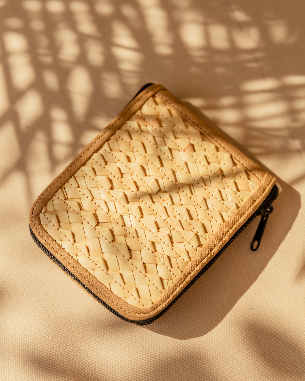Close-up of a woven rattan and canvas zippered pouch on a neutral surface with palm shadow effect.