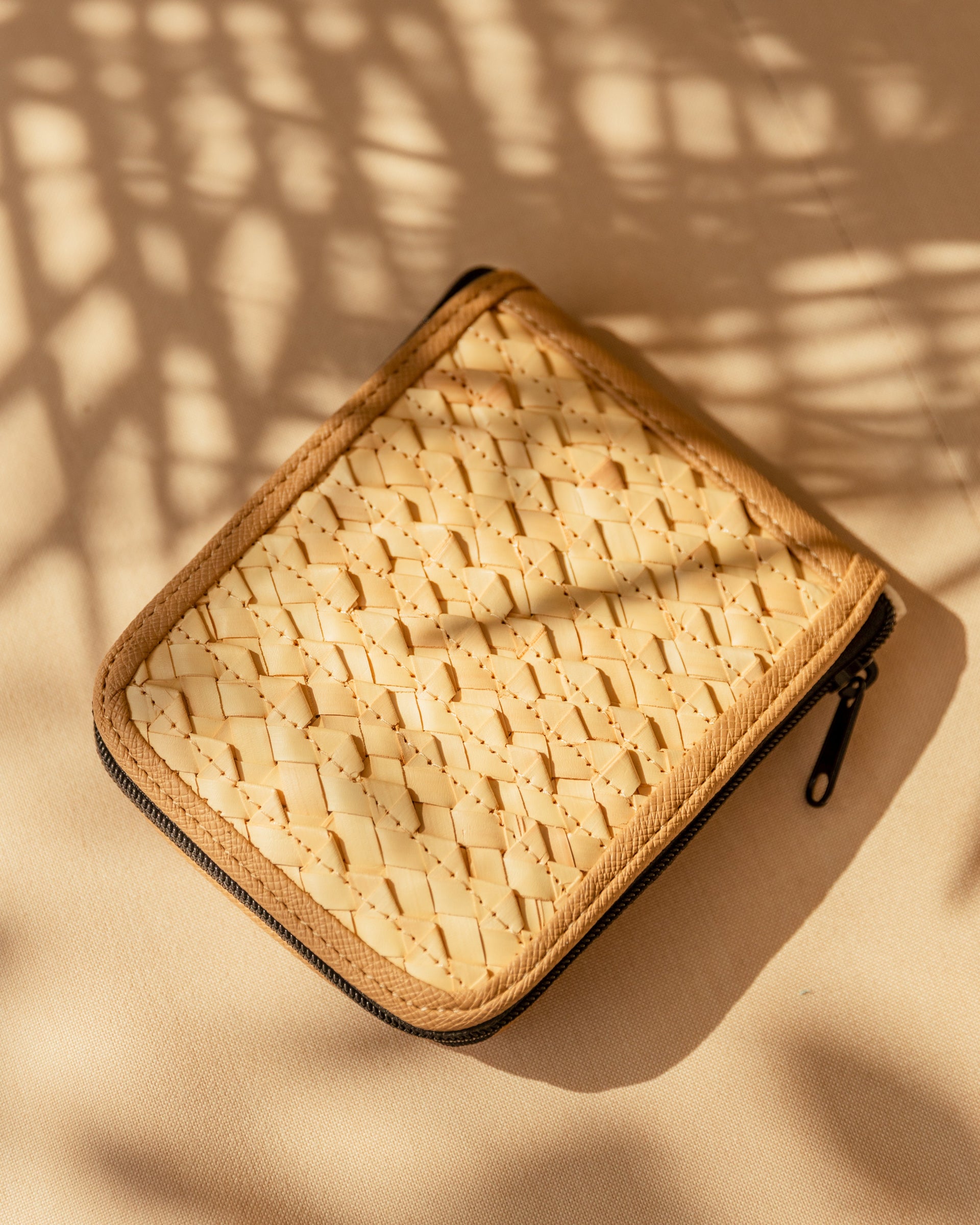 Close-up of a woven rattan and canvas zippered pouch on a neutral surface with palm shadow effect.