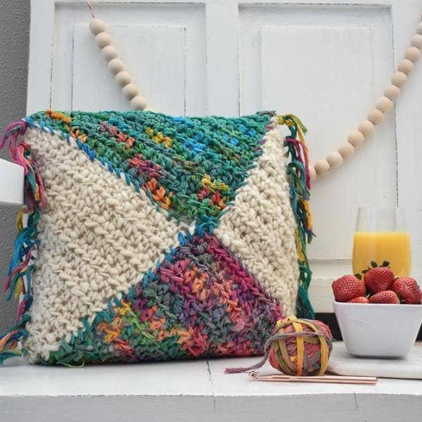 A decorative square throw pillow with fringe and abstract colorful watercolor design sitting on a chair.
