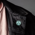 Close up of a circular mint green and chrome enamel pin attached to a black fabric jacket