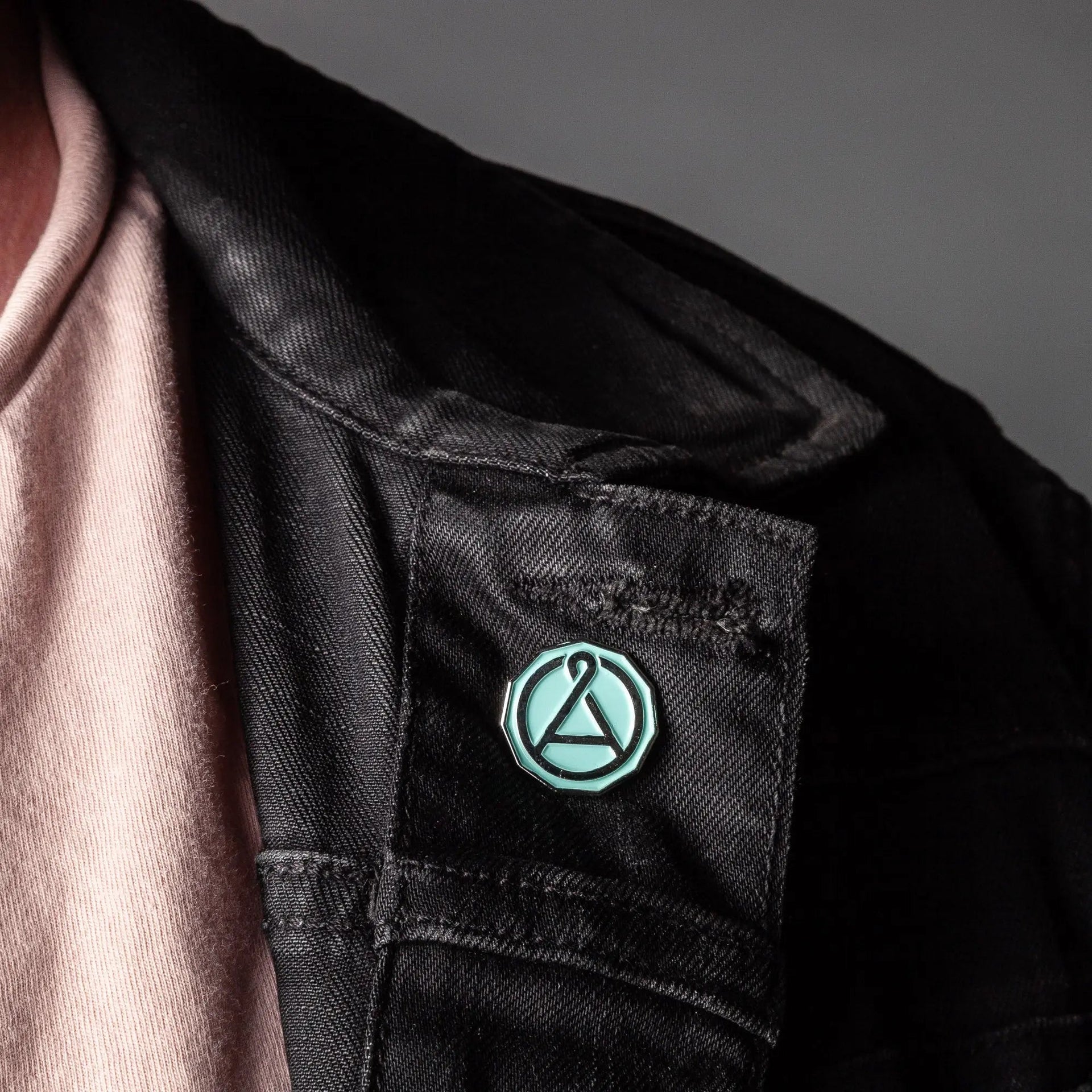 Close up of a circular mint green and chrome enamel pin attached to a black fabric jacket