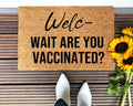Natural brown coir doormat with black text reading 'Wait, Are You Vaccinated?' on a front porch.