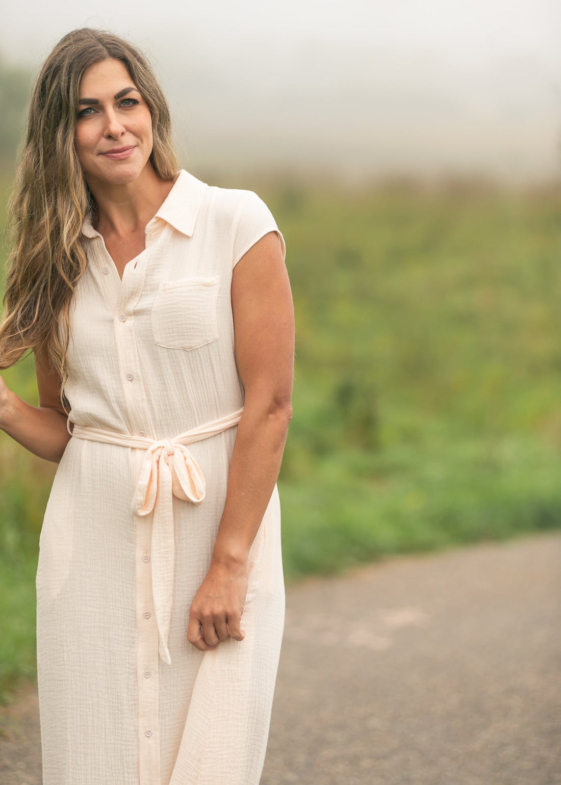 Model wearing a Willow Blush Pink cotton gauze midi shirt dress with cap sleeves and a belted waist standing on a path