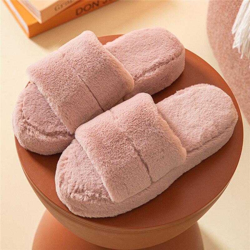 A pair of plush, dusty pink faux fur open-toe slippers on a minimalist bedroom floor.