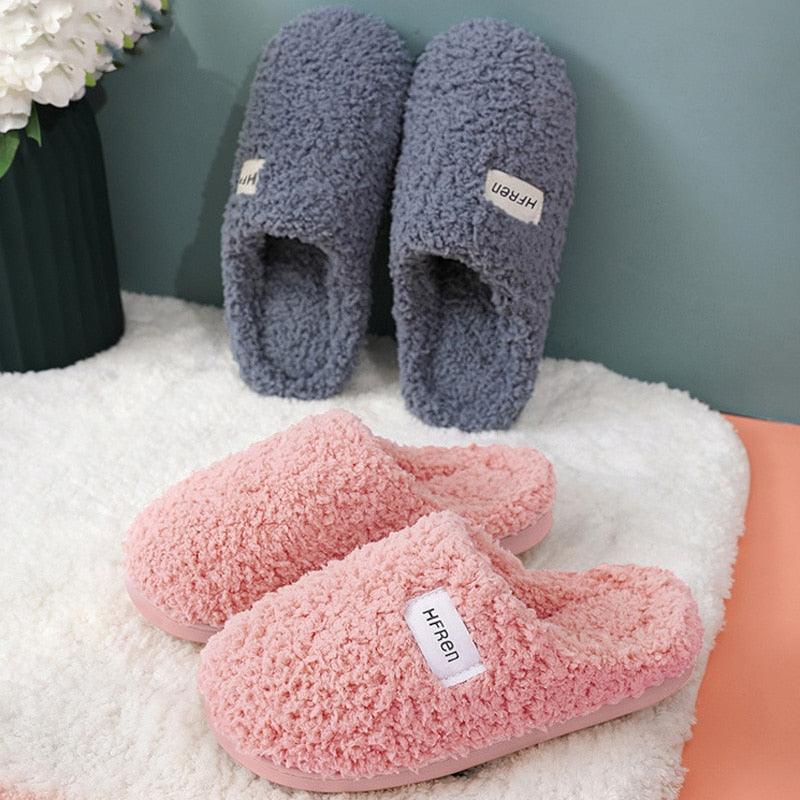 A pair of cozy, minimalist unisex plush slippers in soft fabric on a bedroom floor.
