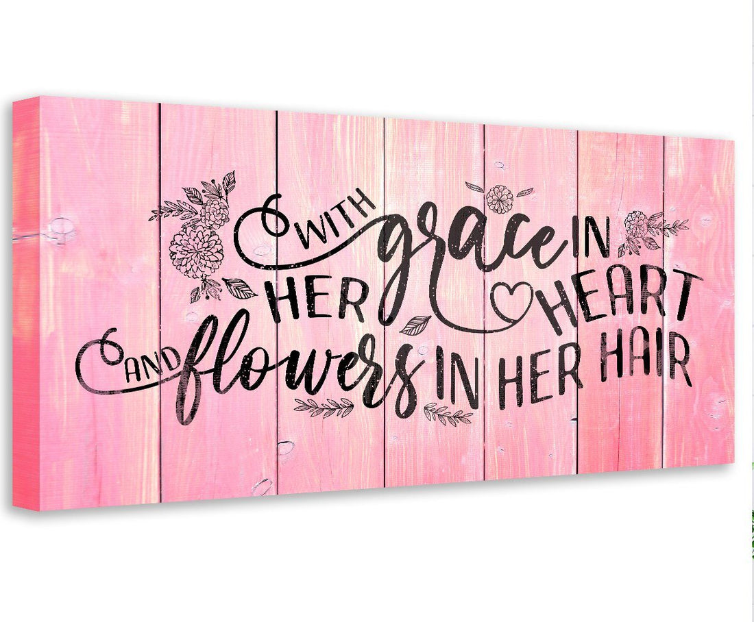 A rectangular canvas wall art piece with a pink faux wood plank background and the black text quote With Grace In Her Heart and Flowers In Her Hair.