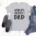 Gray graphic t-shirt with black text reading World's Okayest Dad, laid flat on a wooden surface with shoes and jeans.