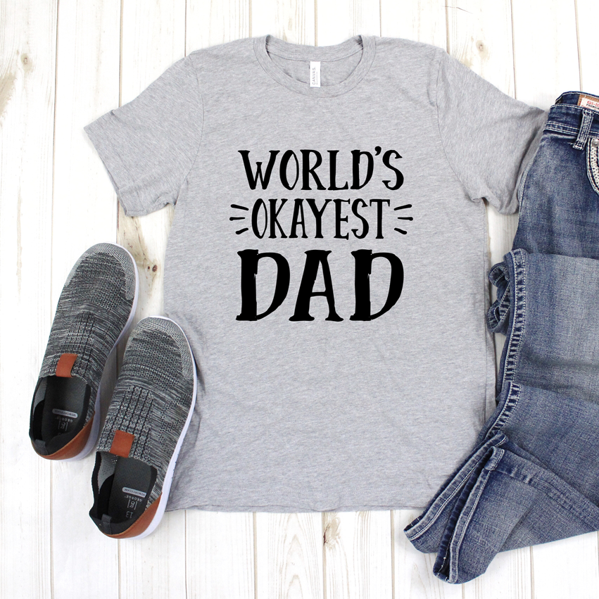 Gray graphic t-shirt with black text reading World's Okayest Dad, laid flat on a wooden surface with shoes and jeans.
