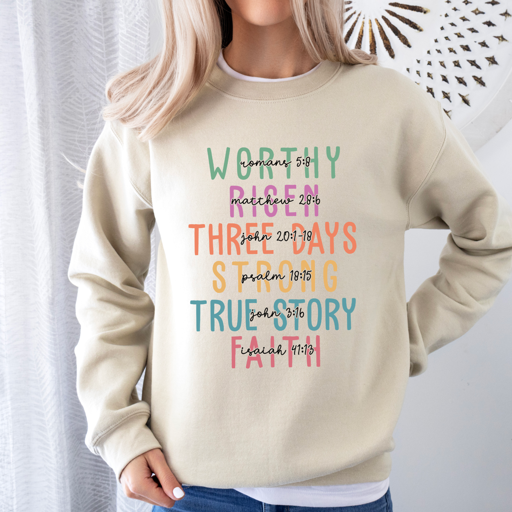 Model wearing a beige crew-neck sweatshirt with colorful Worthy Risen scripture graphic design.