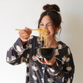 A woman wearing a black bamboo lounge set with a ramen noodle bowl print, featuring a button-up top and matching flared pants.