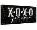 A black and white canvas print displaying XOXO Forever text, hanging above a bed as wall decor.