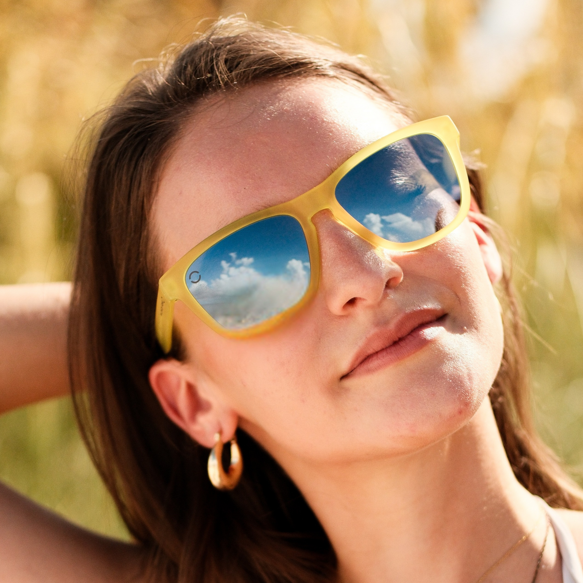 A person wearing bright yellow frame sunglasses with grey polarized lenses against a sunny background