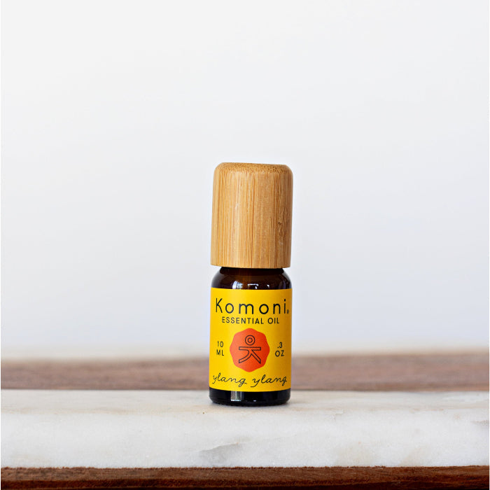 A 10ml amber glass bottle of Ylang Ylang essential oil with a wooden cap next to its yellow packaging box.