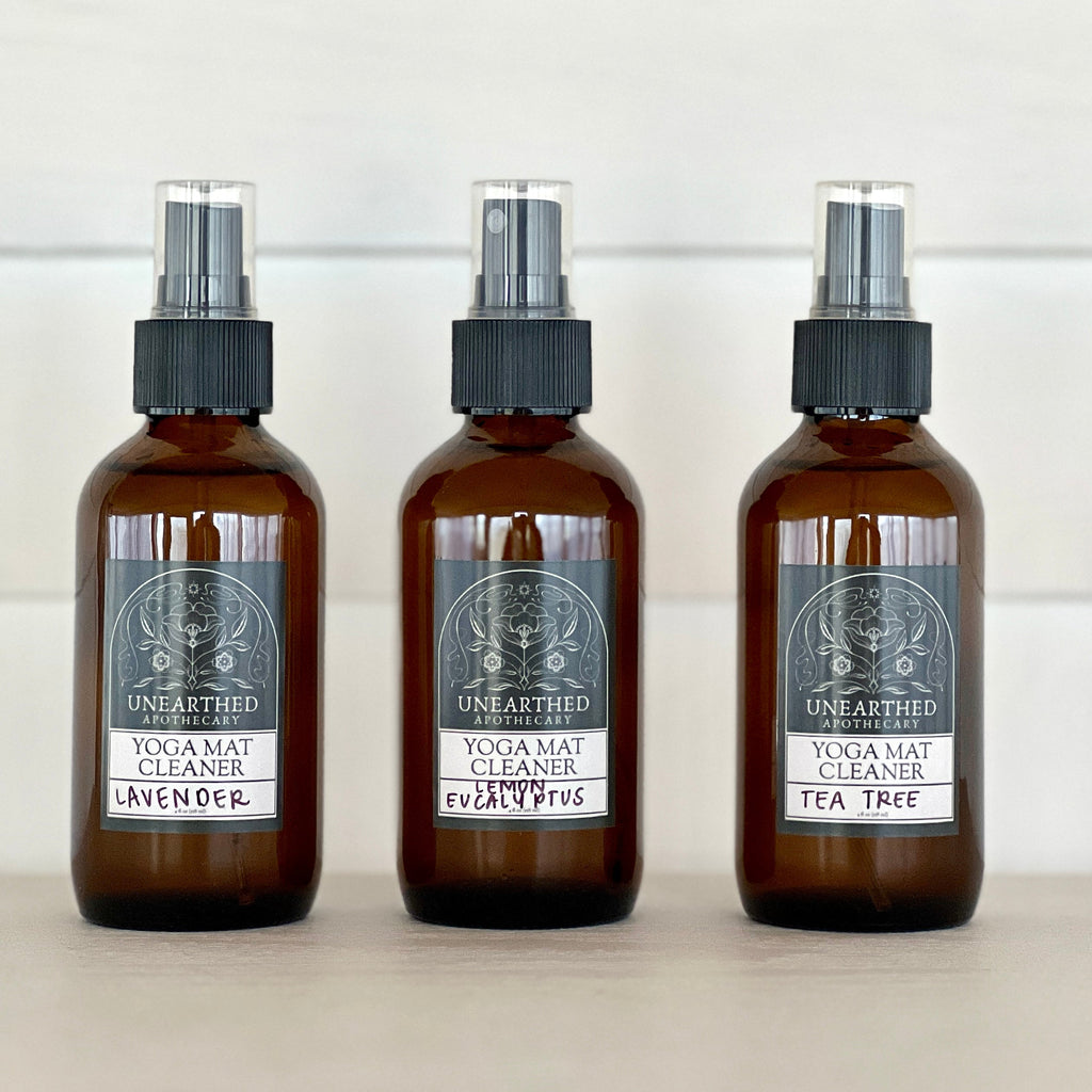 Three glass spray bottles of yoga mat cleaner labeled Lavender, Lemon Eucalyptus, and Tea Tree on a white wooden surface.