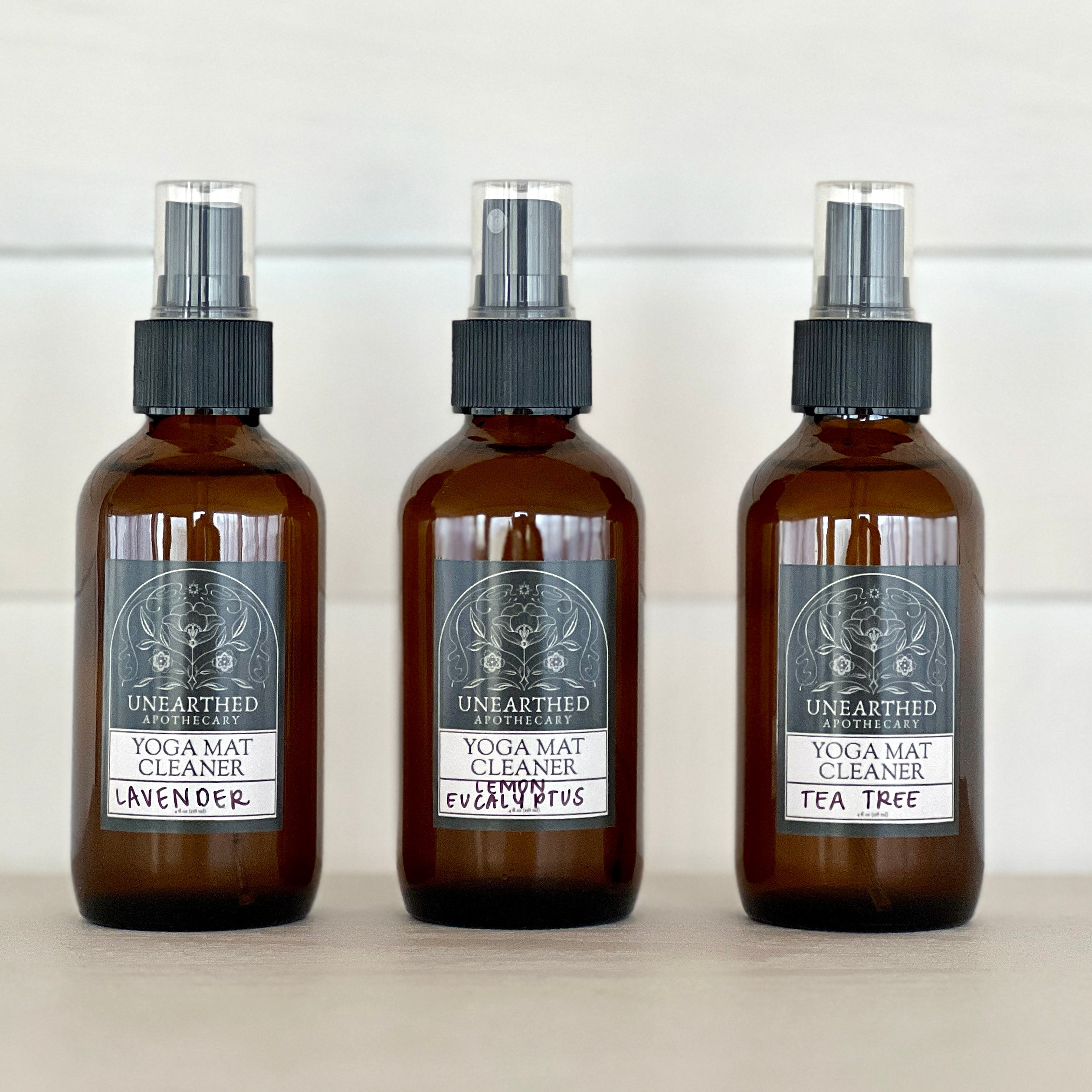 Three glass spray bottles of yoga mat cleaner labeled Lavender, Lemon Eucalyptus, and Tea Tree on a white wooden surface.