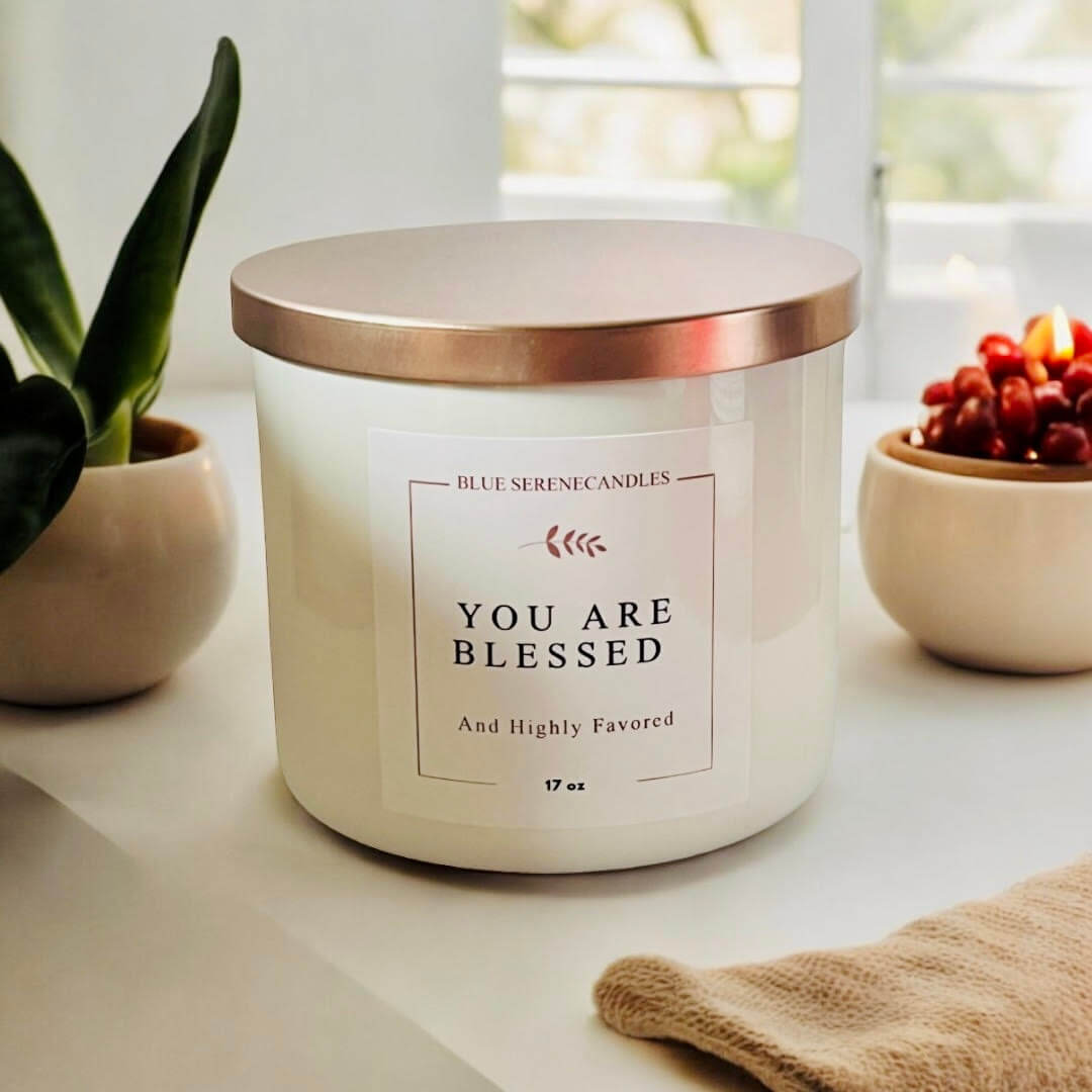 White jar candle labeled You Are Blessed and Highly Favored with a metallic copper lid sitting on a white surface.