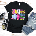 A black short-sleeve unisex t-shirt featuring a colorful graphic that says 'YOU ARE' followed by positive adjectives like kind, brave, smart, and helpful in a doodle art style.